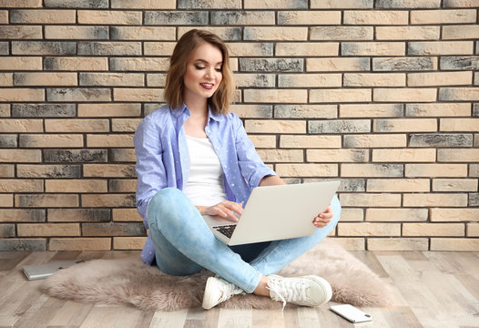 Beautiful young woman using laptop while sitting on floor near brick wall