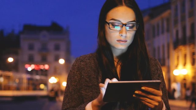 Pretty young woman in glasses walking the city square, scrolling on her tablet computer and looking around in the evening. Close up. Portrait shot. Outdoors