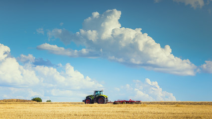 Fototapeta premium In the summer heat of the tractor plows the field after harvest against the bright Sunny sky with clouds.