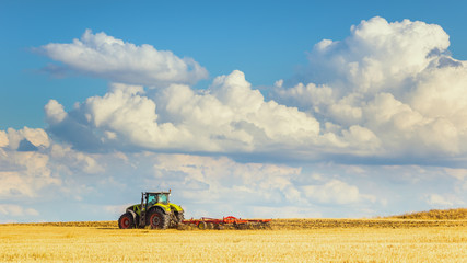 Fototapeta premium In the summer heat of the tractor plows the field after harvest against the bright Sunny sky with clouds.