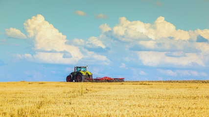 Fototapeta premium In the summer heat of the tractor plows the field after harvest against the bright Sunny sky with clouds.
