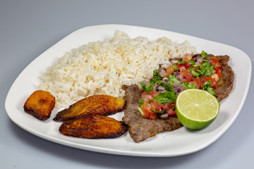 A steak covered in pico de gallo surrounded by plantains and white rice on a white plate. Cuban food.