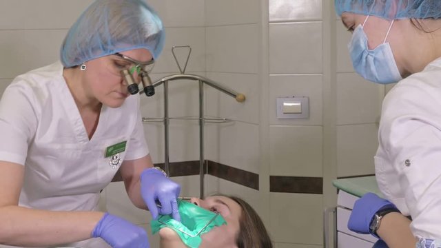 Woman at dentist clinic gets dental treatment to fill a cavity in a tooth. Dental restoration and composite material polymerization with UV light and laser. Installation of dental dam.
