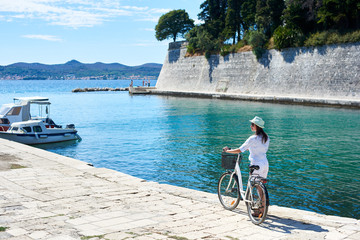 Perfect summer resort landscape. Back view of young woman in sunglasses with backpack riding bicycle along sidewalk by sheltered harbor of resort town with high stone walls. Active lifestyle concept.
