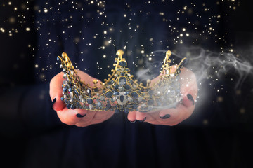 image of lady in black holding queen crown decorated with precious stones and magical glowing mysterious dust. fantasy medieval period. Black queen.