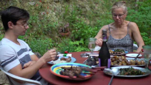 Mature blonde woman talking to her son at a dining table set outside in garden patio.