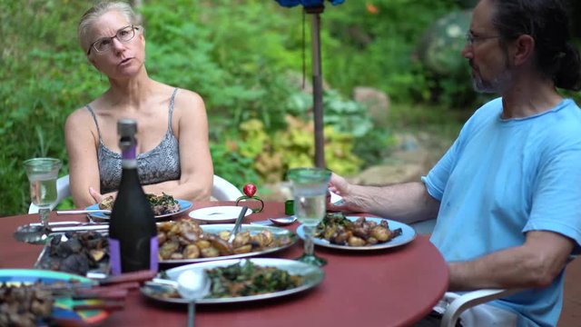 Mature blonde woman talking at the dinner table set outside in patio garden in summer.