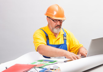 Portrait of an elderly builder in bright working clothes and a construction helmet, which sits at the table with drawings