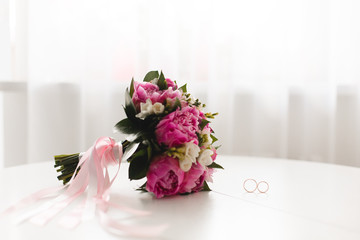 Beautiful pink peonies bouquet  and wedding rings lie on a white table. 