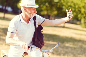 Updating social media. Waist up shot of a stylish elderly gentleman grinning broadly while leaning on his bicycle and taking selfies outdoors.