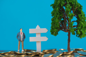 Figure miniature businessman or small people investor standing on coin stack with white wooden board sign and little tree decoration, for money and financial business success concept.