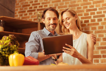 Home day. Attractive young woman keeping smile on her face and holding tablet while standing near her man