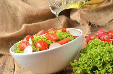 Pouring oil into green salad Lollo Biondo with tomatoes and radishes in a white bowl on wooden table, a pitcher of oil in the background