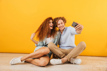 Photo of joyous boyfriend and girlfriend 20s sitting on floor together and taking selfie on smartphone, isolated over yellow background