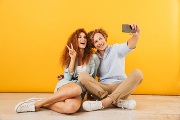Photo of joyful couple handsome man and curly woman 20s sitting on floor together and showing peace sign while taking selfie on smartphone, isolated over yellow background