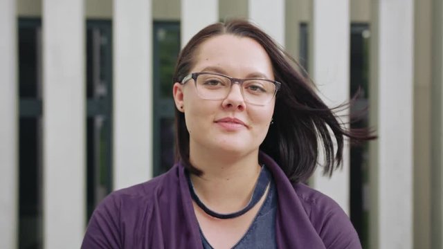 An attractive young lady wearing glasses smiles outdoors. Medium shot