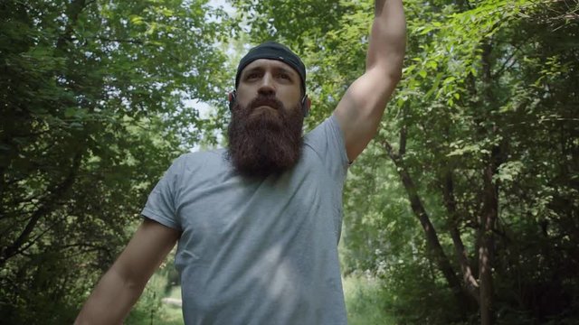 A modern bearded man in a cap trains in the forest and kneads joints. A man prepares to run in the woods and kneads his wrists. Warm up before running. Training in the forest.