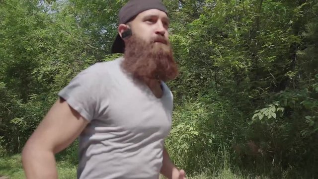 Sport and training: a brutal man with a beard in a sport suit runs through the forest and listens to music. A run in the summer park: a hardy man runs forward. Runner. Cardio workout.