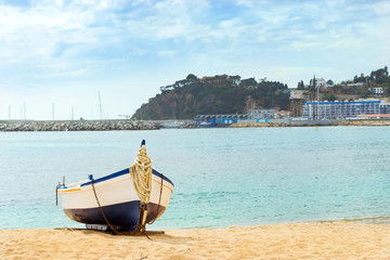 Fishing boat moored on the sandy beach on the background of the seaport. Coast of sandy beach and architecture of Spanish beach resort Blanes in summertime. Costa Brava, Catalonia, Spain