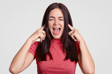 Annoyed brunette woman asks to stop this sound, pluggs ears with both index fingers, exclaims angrily, keeps eyes shut, dressed casually, isolated over white background. Aggressive girl ignores sound