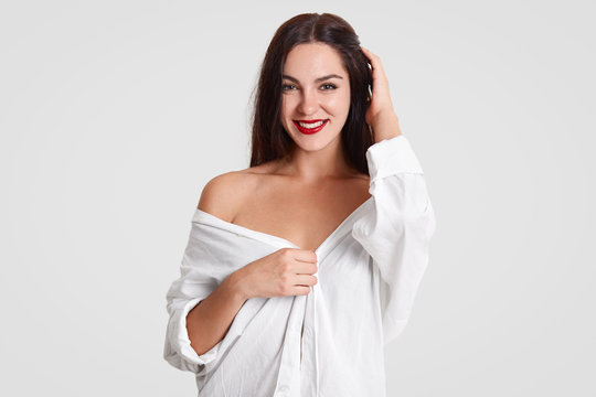 Smiling brunette female with charming look, has make up, healthy skin, red lips, dressed in man`s shirt, shows bare shoulder, isolated over white background. People, beauty and lifestyle concept
