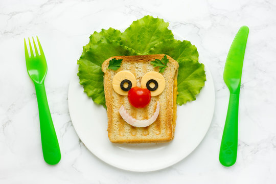 Creative sandwich for kids healthy and funny breakfast. Clown face fried sandwich with green salad, cheese, tomato and ham on white plate top view