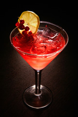 a red cocktail with ice, a slice of lemon and a branch of red currant
