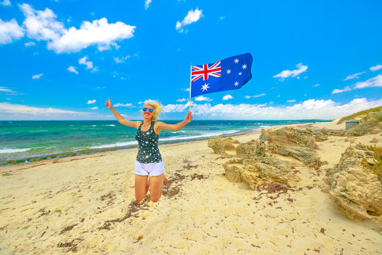 Happy woman jumping on white beach waving Australian Flag. Blonde girl happiness jump in Mettams Pool, Trigg Beach, North Beach neighborhood near Perth, Western Australia. Tourism in Oceania.