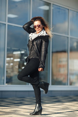 woman in sun glasses a black leather jacket, black jeans posing in front of mirrored windows. Female fashion concept. Outdoor