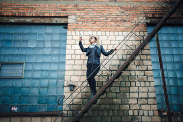 Smiling young woman in sunglasses, a black leather jacket, black jeans standing on an urban metal stair against a brick wall and dances. Woman listening to music on the stairs of industrial building.
