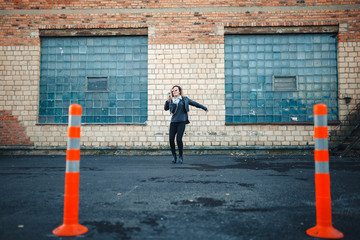 Young beautiful sexy woman with headphones and smartphone in hand in mirrored sunglasses, a black leather jacket, black jeans dancing by the wall of the industrial building of brick on the street