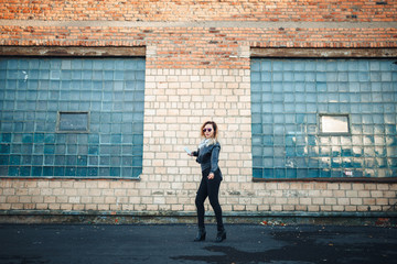 Young beautiful sexy woman with headphones and smartphone in hand in mirrored sunglasses, a black leather jacket, black jeans dancing by the wall of the industrial building of brick on the street