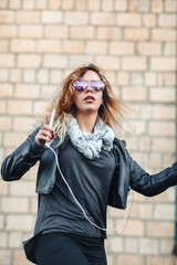 Young beautiful sexy woman with headphones and smartphone in hand in mirrored sunglasses, a black leather jacket, black jeans dancing by the wall of the industrial building of brick on the street