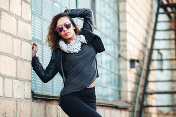 model in sunglasses, black leather jacket, jeans. Posing near a red brick wall