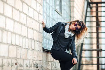 model in sunglasses, black leather jacket, jeans. Posing near a red brick wall