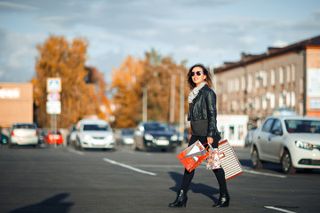 Lovely young woman in sunglasses, a black leather jacket, black jeans turns around holding shopping bags on Parking near the shopping center.