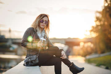 Attractive woman in mirrored sunglasses, a black leather jacket, drinking coffee on the waterfront river in the city in front the setting sun. Waving hair. city,bridge