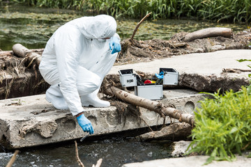 male scientist in protective suit and latex gloves taking sample of water in test flask outdoors