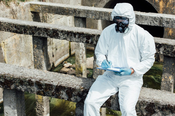  male scientist in protective mask and suit writing in clipboard while sitting near sewerage