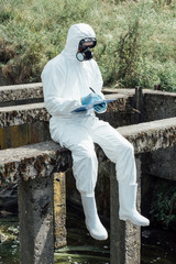 selective focus of male scientist in protective mask and suit writing in clipboard while sitting near sewerage