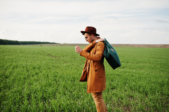 Stylish man in glasses, brown jacket and hat with bag posed on green field.