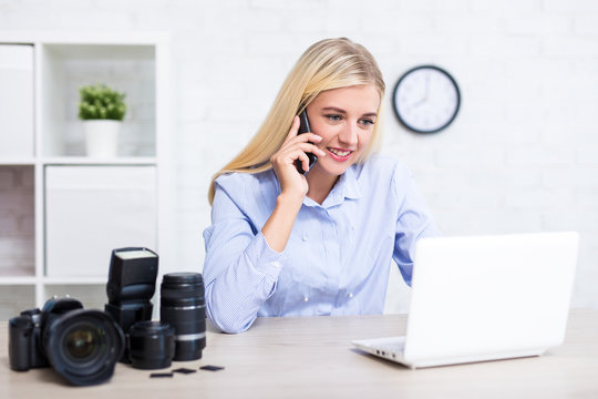 female photographer with camera, computer and photography equipment talking by phone with clients