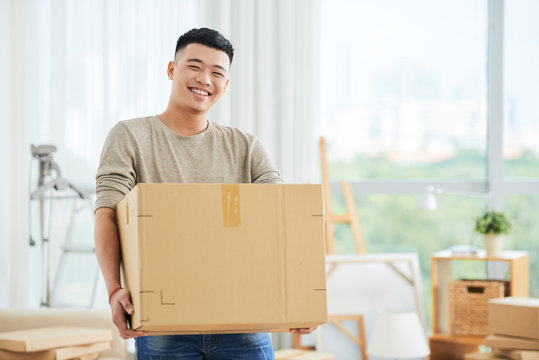 Happy cheerful ethnic male in casual clothes carrying cardboard box and looking at camera on moving day with cartons and drawing easels on blurred background