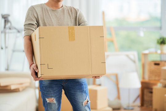 Crop View Of Strong Male In Casual Clothes Carrying Large Cardboard Box On Moving Day With Cartons And Drawing Easels On Blurred Background Of Art Workshop.