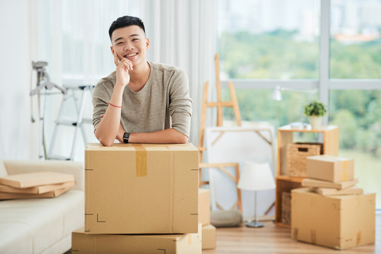 Happy handsome ethnic man in casual clothes standing leaning on pile of large cardboard boxes for moving and looking at camera smiling with finger at cheek on blurred background of packed cartons and