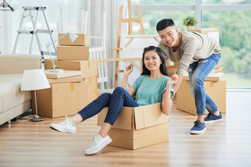 Fototapeta na wymiar Handsome ethnic man pushing large carton box with sitting young beautiful woman on moving day, both looking at camera and smiling