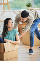 Beautiful young Asian woman in casual clothes sitting in large cardboard box and looking happily at handsome man pushing carton on blurred background of new apartment.