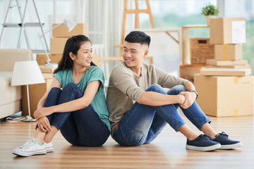 Happy Asian man and woman sitting together on floor of new apartment on moving day and looking at each other smiling with packed carton boxes on blurred background .