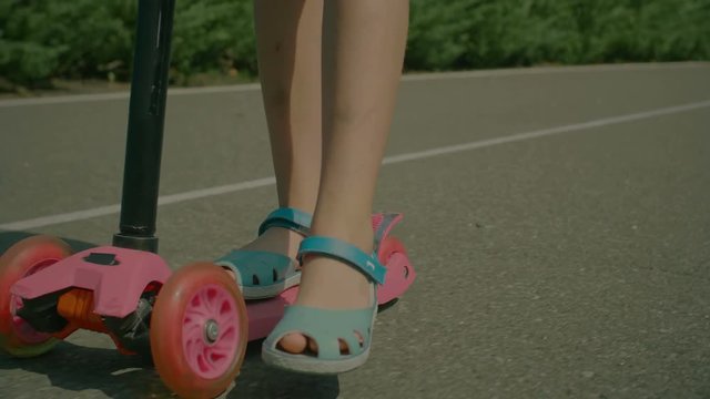 Closeup of feet of little girl in rubber sandals pushing while riding kick scooter in public park. Low angle view. Joyful child enjoying her ride on tricycle scooter outdoors. Slow motion.