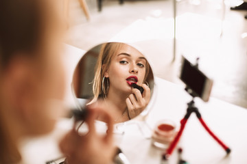 Young pretty girl looking in little mirror on table using red lipstick while recording new video...